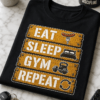Majica z napisom EAT SLEEP GYM REPEAT na lesenih tablah z ikonami hrane, postelje, uteži in puščic
